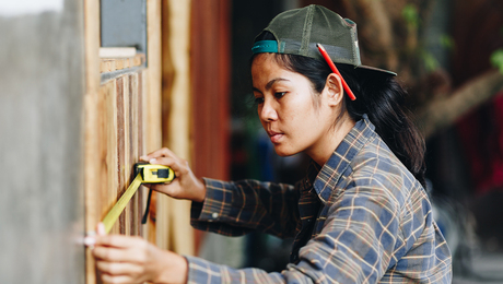 Woman wearing a hat with a pencil behind her ear measures a wall using a tape measurer.